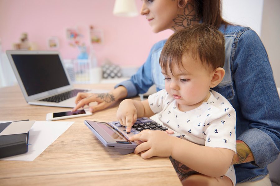 Single parent reviewing loan documents on a phone while sitting at a kitchen table with bills and a child nearby.